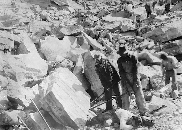 Posed photograph by the American Red Cross of a quarry rescue: men with iron bars prying a rock off a man